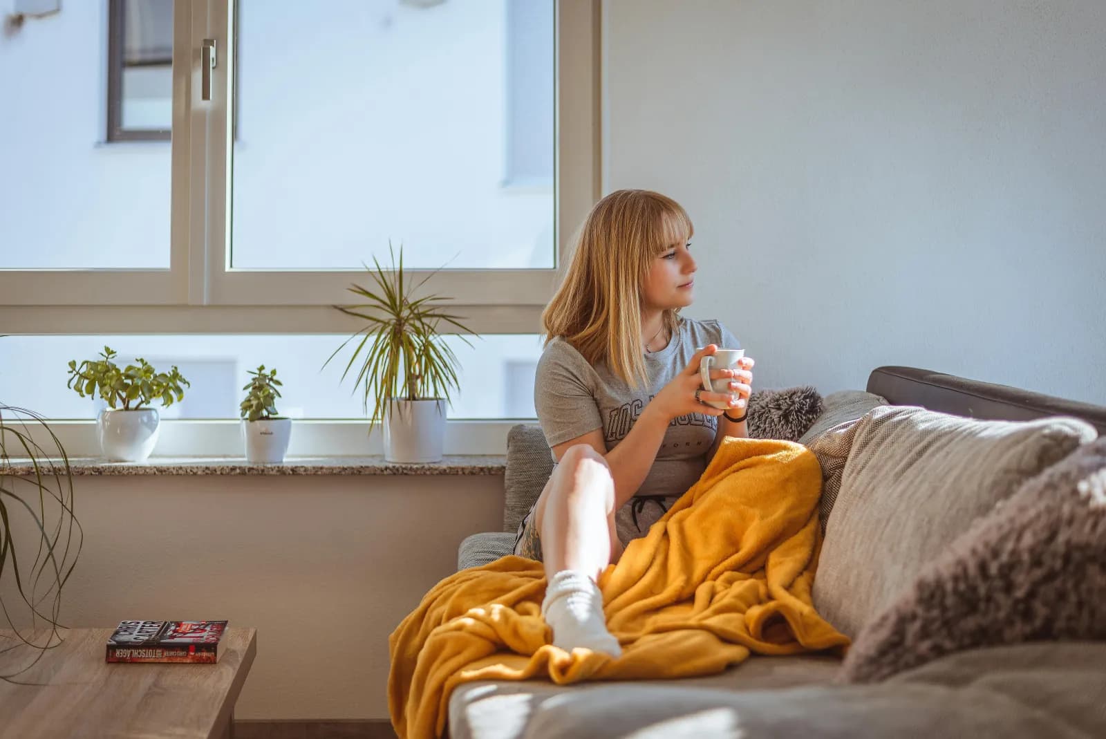 Ung kvinne med kaffekopp i en sofa ved vinduet med pledd og planter